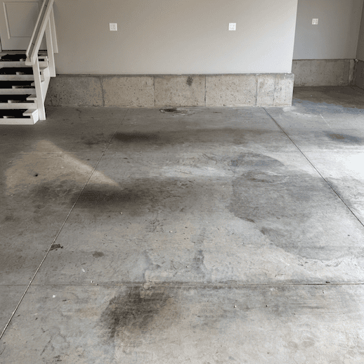 Stained bare concrete garage floor with dark blotches and a staircase in the corner before coating