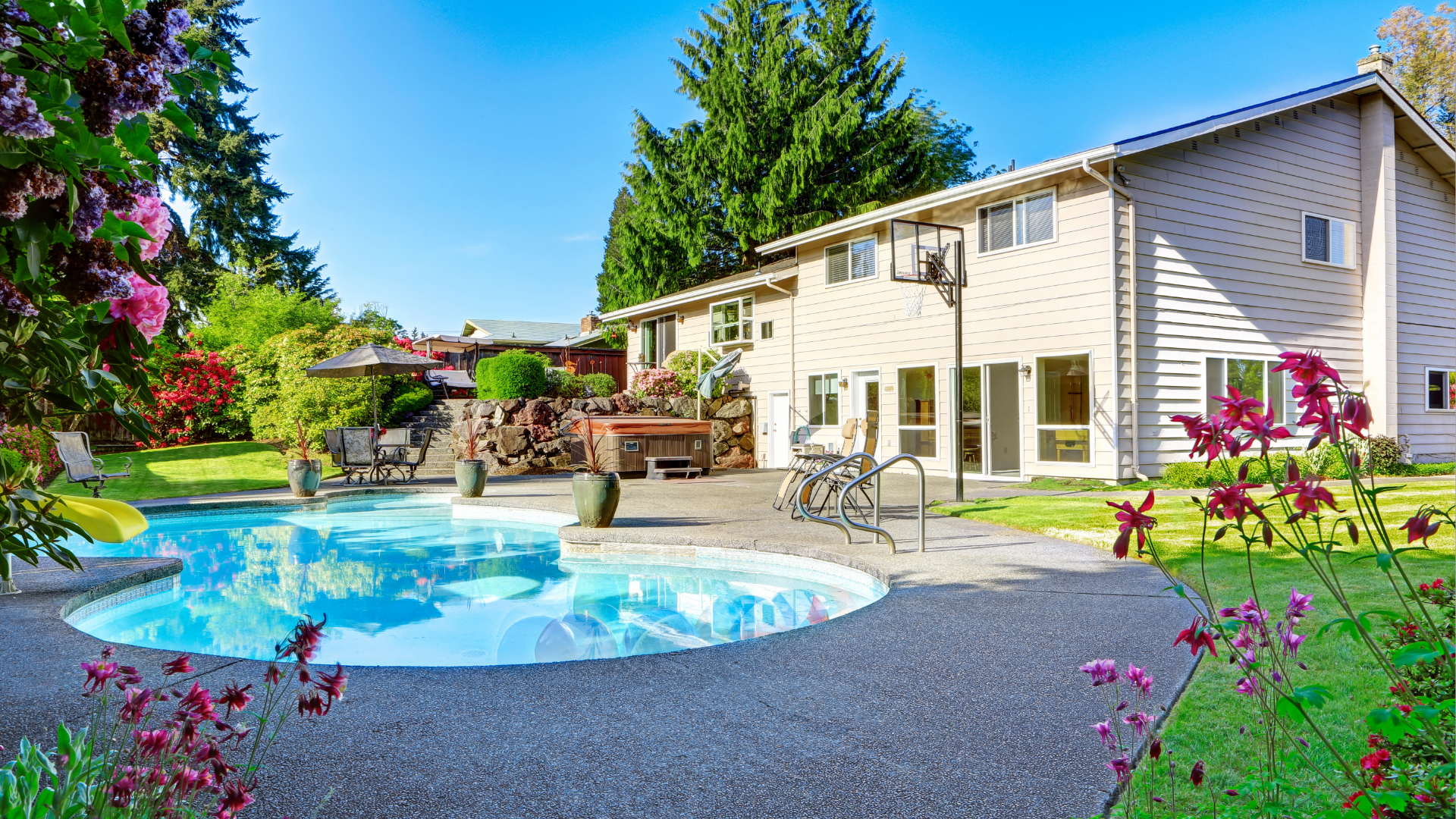 Two-story beige house with a backyard pool, gray concrete deck, lawn and flowers